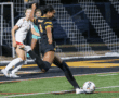 Soccer player in black and yellow uniform kicks ball towards goal during a match, opposing team watches intently.