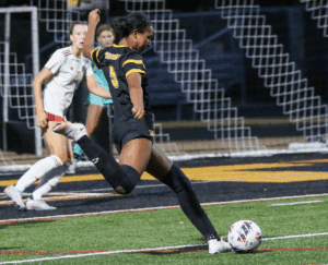Soccer player in black and yellow uniform kicks ball towards goal during a match, opposing team watches intently.