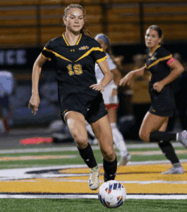 Soccer player in black and yellow uniform dribbles ball during a game on a green field.