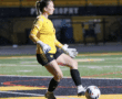 Soccer goalie in yellow jersey kicking a ball on a rainy field at night.