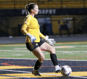 Soccer goalie in yellow jersey kicking a ball on a rainy field at night.