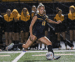 Soccer player in blue uniform kicks ball on rainy field during game, with team in yellow jackets in background.