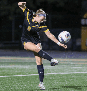 Soccer player in action kicking ball during night match, wearing black and yellow uniform.