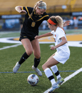 Two female soccer players compete for the ball on a grassy field during a match.