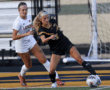 Two female soccer players in action on the field, one in black and one in white uniforms, near the goal line.