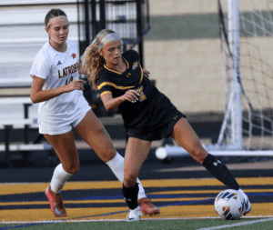 Two female soccer players in action on the field, one in black and one in white uniforms, near the goal line.