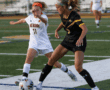 Two female soccer players compete for the ball on a field during a match, wearing white and black uniforms.