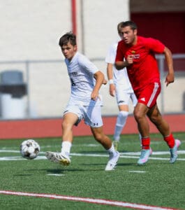 Soccer players in action during a match, one in white kicking the ball, another in red chasing on a green field.