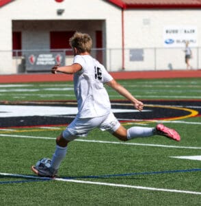 Soccer player in a white uniform kicking a ball on a grass field during a match.