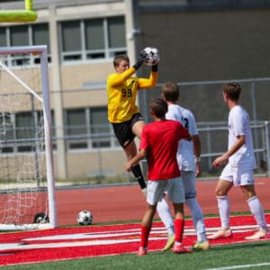 Goalkeeper in yellow jersey catches soccer ball mid-air during a game on a sunny day.