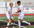 Two soccer players celebrate on the field, wearing white uniforms, with stadium seats in the background.