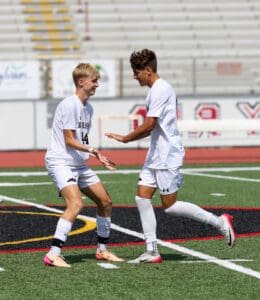 Two soccer players celebrate on the field, wearing white uniforms, with stadium seats in the background.