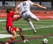 Soccer players battling for the ball on a sunny field, wearing red and white jerseys.