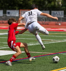 Soccer players battling for the ball on a sunny field, wearing red and white jerseys.