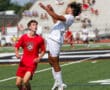 Soccer match action: player in white jersey heads ball while player in red watches on a sunny day.