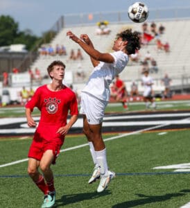 Soccer match action: player in white jersey heads ball while player in red watches on a sunny day.