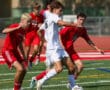 Soccer players in action during a competitive match on a sunny day, wearing white and red jerseys on a green field.