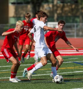Soccer players in action during a competitive match on a sunny day, wearing white and red jerseys on a green field.