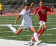 Soccer player in white uniform kicks ball, pursued by opponent in red, during an intense match on a sunny day.