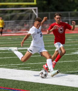 Soccer player in white uniform kicks ball, pursued by opponent in red, during an intense match on a sunny day.