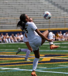 Soccer player in white jersey skillfully controls the ball mid-air during a match on a sunny day.