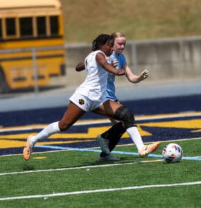 Two female soccer players compete for the ball on a sunny field, one wearing a knee brace.