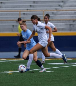 Soccer players in action on a field during a match, one in white, another in blue, competing for the ball.