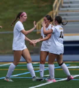 Three female soccer players in white jerseys celebrating on the field with high-fives.