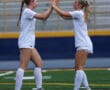 Two female soccer players in white uniforms high-five on the field.