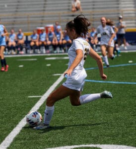 Soccer player in white uniform kicks ball on field during a game, with teammates and opponents in the background.