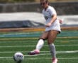 Female soccer player in white uniform kicking a ball on a green field during a game.