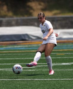 Female soccer player in white uniform kicking a ball on a green field during a game.