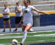 Soccer player in white uniform kicks ball on field during a match, with blurred spectators in background.