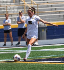 Soccer player in white uniform kicks ball on field during a match, with blurred spectators in background.