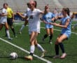 Female soccer match on a sunny day; player in white controls the ball while blue team defends.