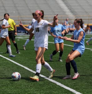 Female soccer match on a sunny day; player in white controls the ball while blue team defends.