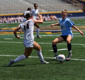 Two female soccer players compete for the ball on an outdoor field during a match.