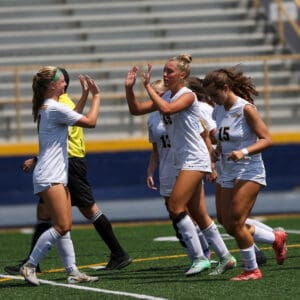 Soccer team celebrates with a high-five on the field during a game under sunny skies.