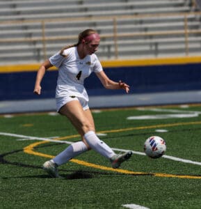 Soccer player in action, kicking ball on field during a sunny day match, wearing white uniform and number 4.