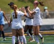 Soccer players in white uniforms celebrating with high-fives on the field during a match in sunny weather.