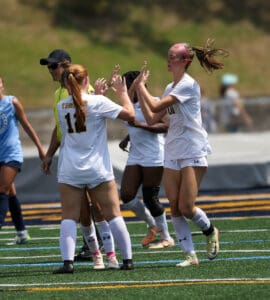 Soccer players in white uniforms celebrating with high-fives on the field during a match in sunny weather.