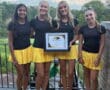 Four young women in matching yellow skirts hold a first place award outdoors on a sunny day.