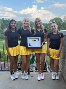 Four young women in matching yellow skirts hold a first place award outdoors on a sunny day.