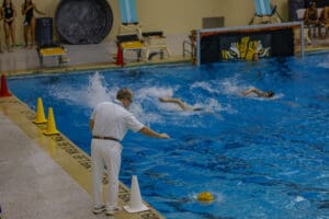 Swimming competition with referee and swimmers racing in pool. Water splash and yellow ball visible.