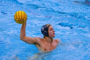 Water polo player in action holding a yellow ball, wearing a numbered cap in a pool.
