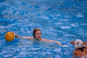 Water polo players in action, one holding a yellow ball, wearing helmets, competing in a pool match.