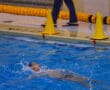 Swimmer practicing backstroke in indoor pool, focused on technique amidst splashing water.