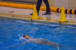 Swimmer practicing backstroke in indoor pool, focused on technique amidst splashing water.