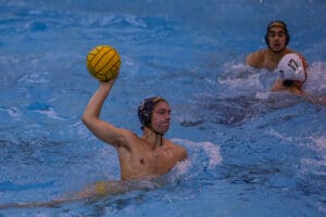 Water polo player poised to throw a yellow ball during a competitive match in a swimming pool.