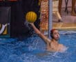 Water polo player prepares to throw a ball by the goal during a match, wearing a helmet and in action mode.